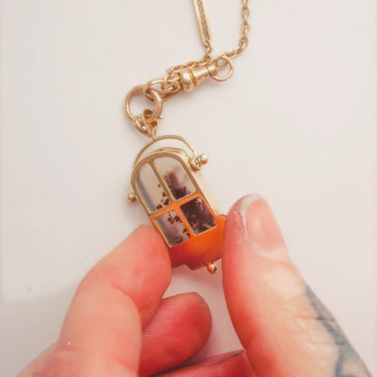 Close-up of a hand holding a gold pendant shaped like a small window with a scenic image behind frosted glass on a chain