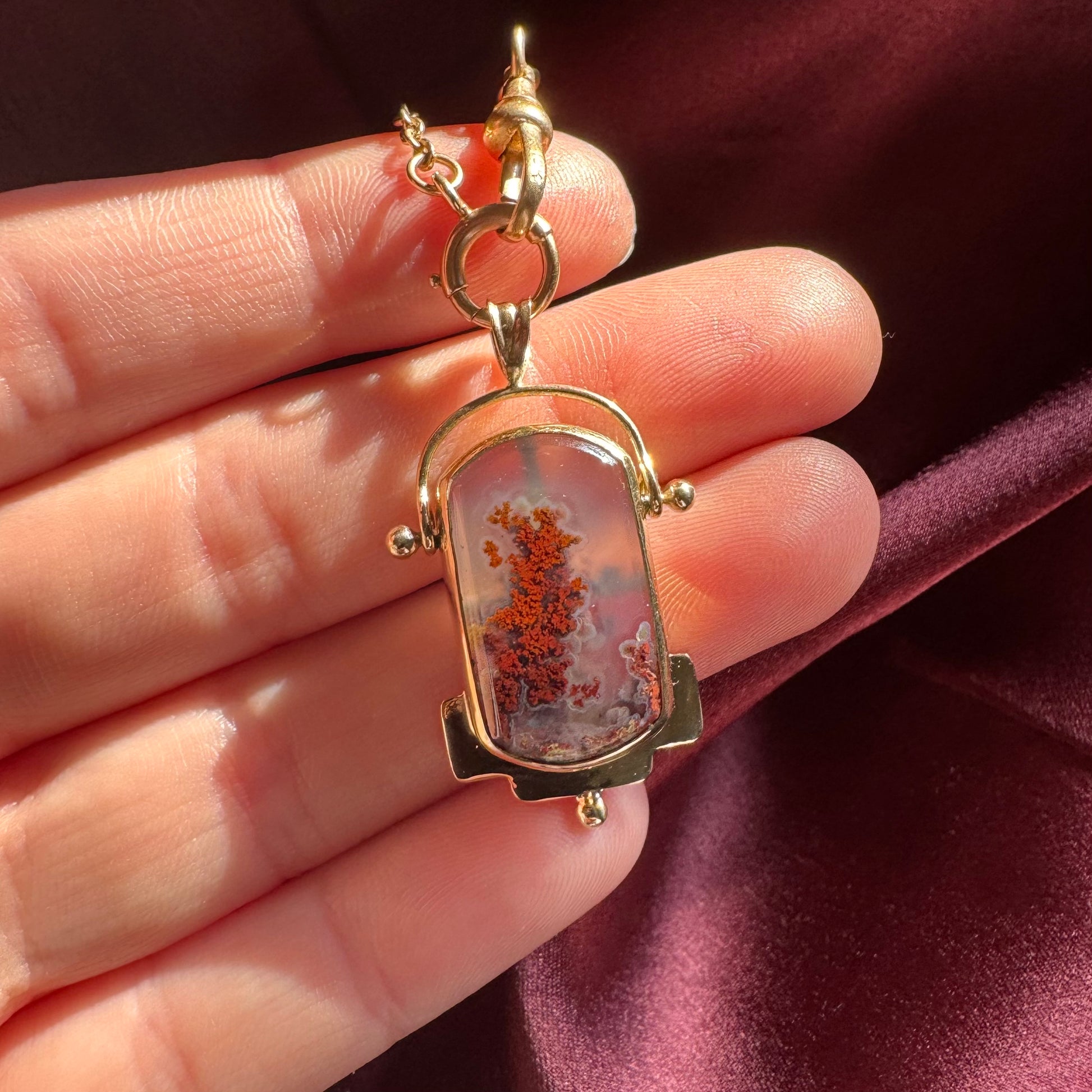 Gold pendant with a central stone held between fingers against a dark maroon background