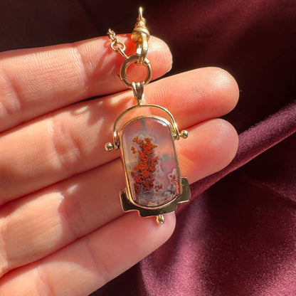 Gold pendant with a central stone held between fingers against a dark maroon background