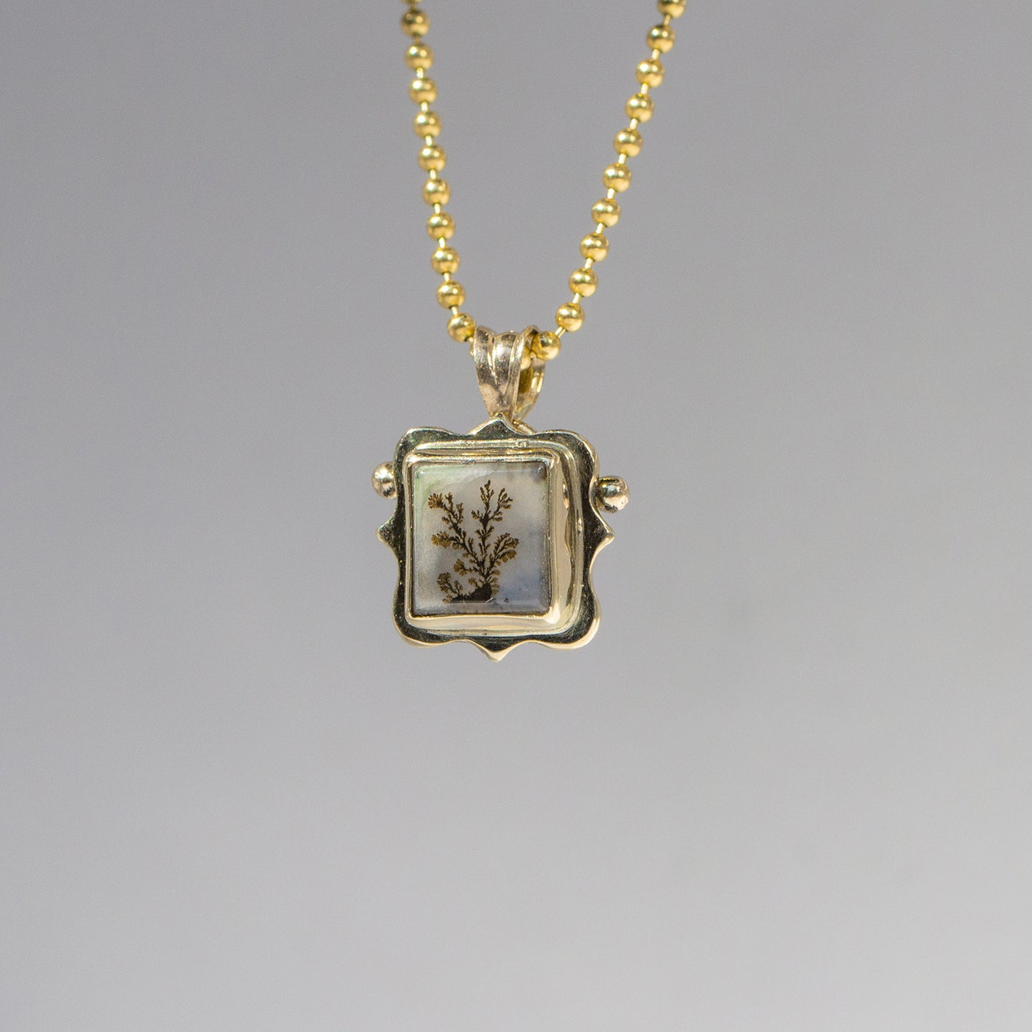 Gold necklace with a square pendant featuring a botanical design on a gray background