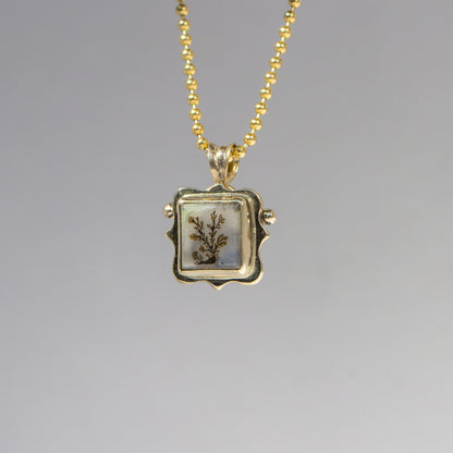 Gold necklace with a square pendant featuring a botanical design on a gray background